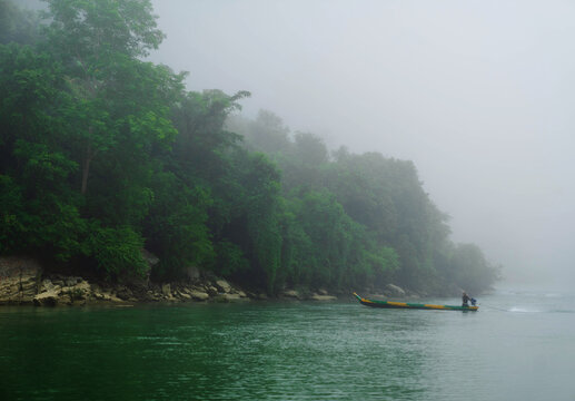 View of a small boat glides through the emerald water, past the verdant forest shrouded in ethereal mist, Bandarban, Chittagong Division, Bangladesh.