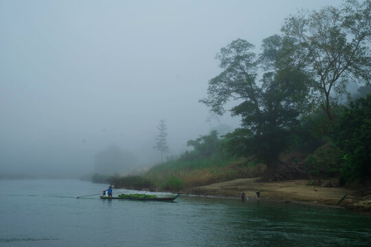 View of a lone boatman rows silently through the misty waters, the riverbank blurred with fog, Bandarban, Chittagong Division, Bangladesh.