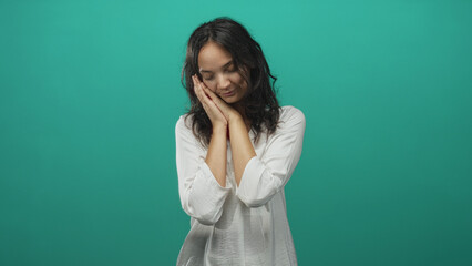 Obraz premium Young hispanic woman resting hands against cheek in a teal studio with white blouse and soft lighting, gentle pose and closed eyes; peaceful rest.