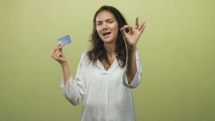 Young hispanic woman holding blue creditcard, fingers forming ok sign in studio backdrop; approval trust.