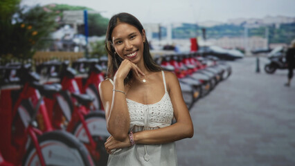 Fototapeta premium Thai woman posing with hand under chin by a row of red rental bicycles at a street bike dock, smiling and turned slightly;contentment.