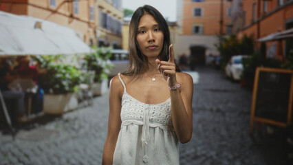 Fototapeta premium Thai woman in white sundress shows bare shoulder and points index finger to temple on a cobbled street lined with cafes; idea thinking confidence.