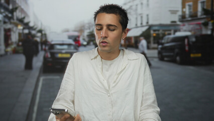 Fototapeta premium Young man talking on smartphone in urban city street, wearing white shirt, surrounded by vehicles and buildings, illustrating communication and modern lifestyle.