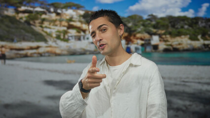 Fototapeta premium Young man in white shirt gesturing expressively at a sandy beach with clear blue sky and rocky background, capturing a lively seaside moment.