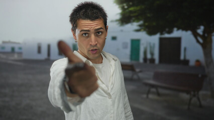 Fototapeta premium Young hispanic man wearing loose white shirt points finger to camera on street showing hoop earrings; anger frustration tension confrontation.