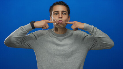 Naklejka premium Young hispanic man making funny face in grey sweater against isolated blue background wall with fingers pointing at cheeks for humorous expression