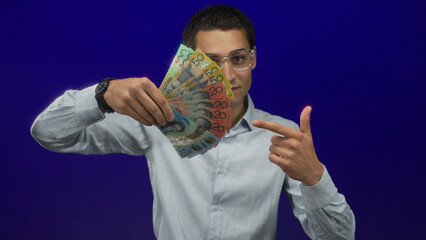 Young hispanic man holding australian money against an isolated blue background, showcasing...
