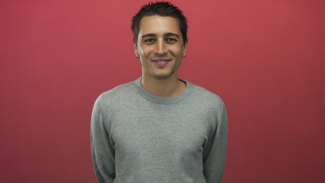 Hispanic man smiling confidently in front of an isolated red background wall, dressed in a casual grey sweater while posing with a friendly demeanor.