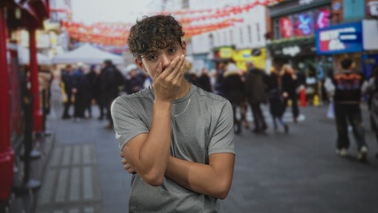 Fototapeta premium Teenage boy rests chin on fist with crossed arms in a crowded street with lanterns, storefronts and a market tent; thoughtful confidence.