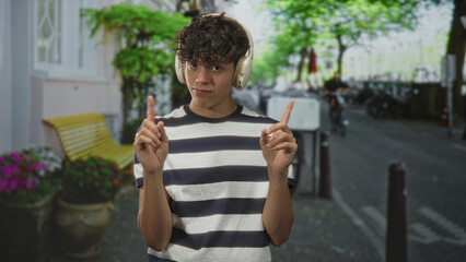 Fototapeta premium Man wearing headphones points both index fingers while standing on a street by a bench with potted flowers; playful listening.