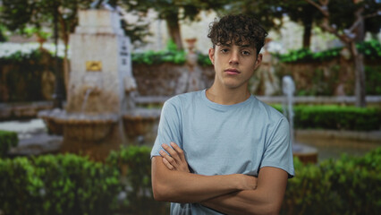 Teen boy in a blue tshirt standing with arms crossed and direct gaze by a stone fountain in a...