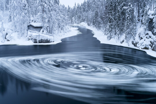 View of swirling, hypnotic vortex carves through the icy river, framed by snow-laden trees and a quaint cabin, a winter's tale unfolds, Myllykoski, Finland.