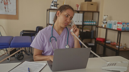 Obraz premium Young woman doctor wearing scrubs and stethoscope speaking on phone at clinic desk, typing on laptop with patient forms and medical shelves visible; stress.