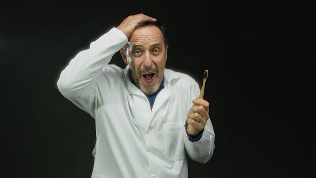 Man in doctor coat holding bamboo toothbrush against black background expresses surprise with hand gestures.