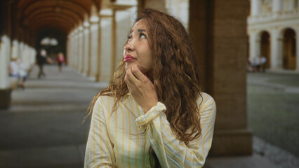 Fototapeta premium Woman touching chin with frown and hand near mouth on historic street arcade, striped yellow shirt visible; quiet concern.
