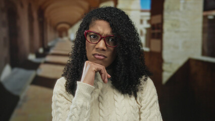 Fototapeta premium Woman with curly hair wearing cream sweater and red glasses scratches head next to building facade; confusion doubt hesitation uncertainty.