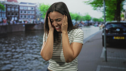 Fototapeta premium Woman covering face with hands on a street canal beside parked cars and buildings, striped shirt visible; vulnerability distress resilience.