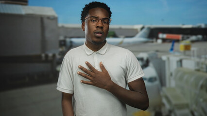 Man hand on chest stands at airport terminal outdoors by parked plane under bright blue sky;...