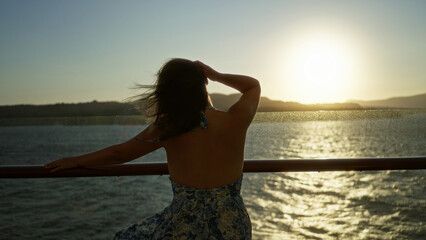 Woman with bare back leaning on ship rail at deck watching the golden sunset, dressed in floral halter dress with hair blowing in wind; quiet serenity.