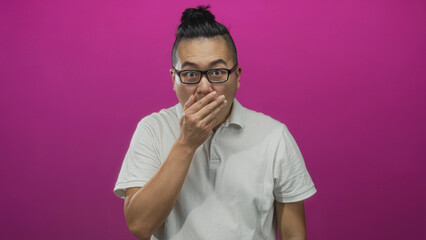 Fototapeta premium Young chinese man wearing glasses covering mouth with hand in studio with magenta backdrop; surprise secret reaction.