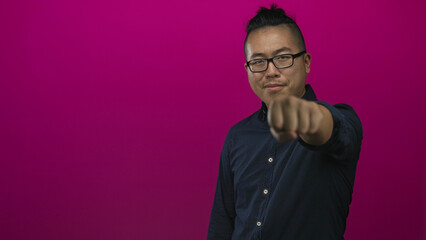 Fototapeta premium Man wearing glasses and navy shirt with fist extended toward camera punching gesture in magenta studio; confidence empowerment resilience.