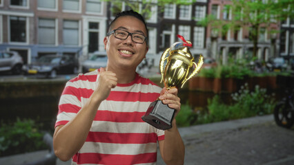 Man holding trophy and making a fist pump on street by canal with parked cars and row houses; pride...