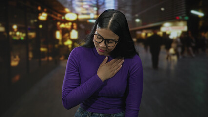 Obraz premium Young latin woman clutching chest on urban street at night with blurred storefronts and neon lights; gratitude.