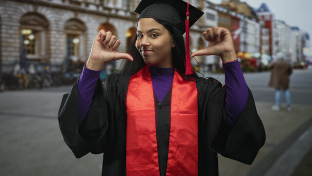 Woman in black graduation gown and mortarboard gestures thumbs down with both hands on busy city street; disappointment.