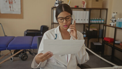 Woman doctor in white coat holds phone to ear with hand while reading a medical report at a clinic...