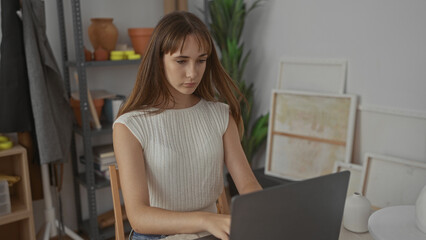 Woman typing on laptop in artisan studio with hands on keyboard and canvases and pottery visible; focused creativity.