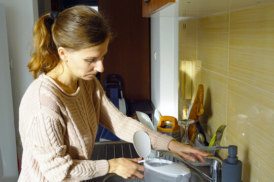 A woman fills a jug with water for filtering. Illustrates predictable home micro-actions, routine life support, slow domestic task sequence, steady maintenance rhythm, and daily self-care ritual.
