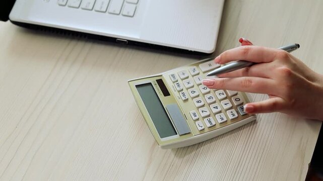 Female hands with manicured nails are using a calculator and a pen on a desk, with a laptop in the background, performing accounting and finance tasks

