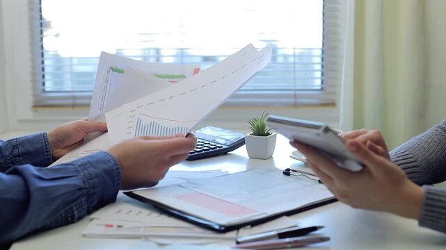 Professional businesswomen collaborating at desk, analyzing financial reports using calculator, focusing on data-driven strategic financial planning and corporate performance evaluation
