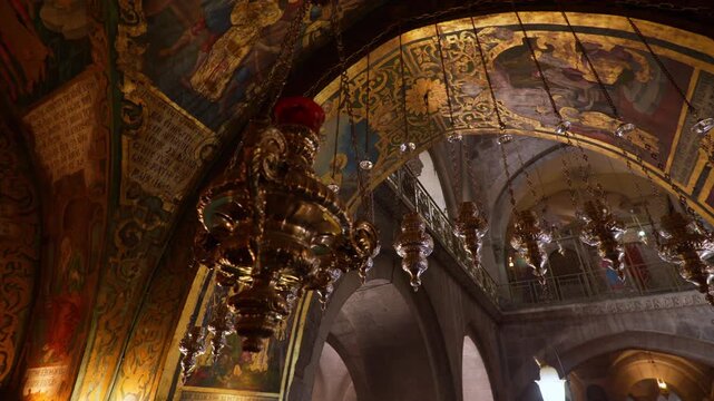 Moving panoramic shot of gilded hanging lamps and Byzantine mosaics beneath stone arches inside the Church of the Holy Sepulchre in Jerusalem, Israel. Sacred Christian interior with warm light