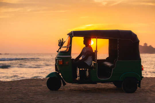 Local tuk tuk driver waiting inside vehicle on sandy beach during ocean sunset in Sri Lanka. Warm evening light and calm tropical atmosphere.