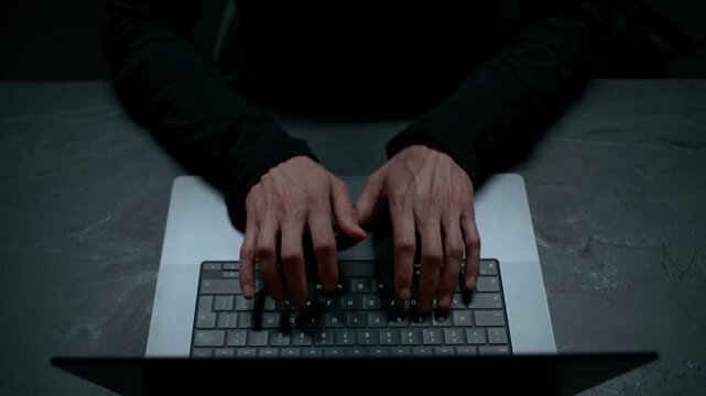 Hacker in a Black Hoodie Breaking Code in a Dark Room &ndash; Crane Shot from a Close-Up of Fingers Tapping on the Keyboard to a View of the Person&rsquo;s Silhouette from Behind the Laptop