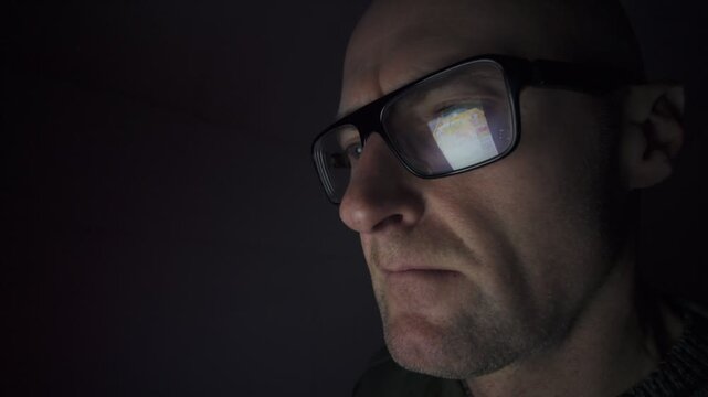 CU Man sat in dark room using a computer, refection in his glasses. Hacker,  social media scrolling. Male mental health.