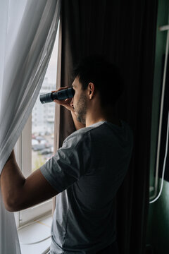 Man curiously peering out window using monocular, watching with intense emotion. Engaging in voyeurism. Observing surveillance. Peeping through curtains. Maintaining vigilance.