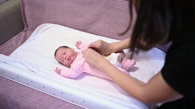 Caring mother gently dressing her adorable newborn daughter in a pink onesie. The infant is lying on a soft changing mat while the parent carefully buttons up the cozy pajamas after changing a diaper