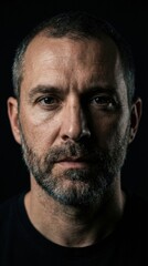Obraz premium Handsome mature man with a salt-and-pepper beard and intense gaze poses for a dramatic, moody headshot portrait on a solid black background