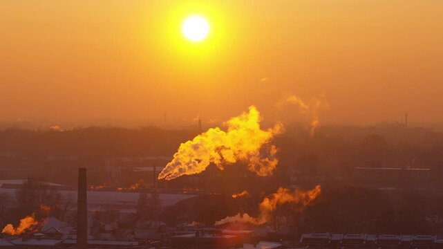 Aerial sunset over Riga, Latvia, shows tall chimneys and smokestacks. Backlit steam and smoke billow over snow roofs as the sun sinks, with warm saturated light.