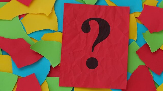 A red note paper with a question mark on it sitting atop a colourful collection of torn paper pieces. Close up.