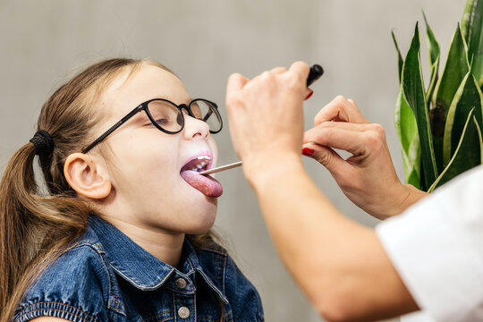 Young girl with glasses undergoing a throat examination by a healthcare professional using a tongue depressor in a clinical setting with a plant in the background
