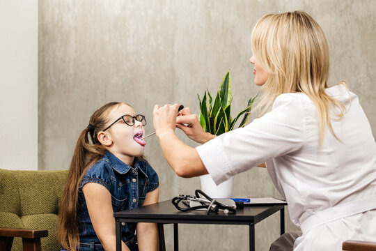 Female pediatrician examines girl with glasses using tongue depressor in a modern clinic setting with green plant and medical equipment on the table