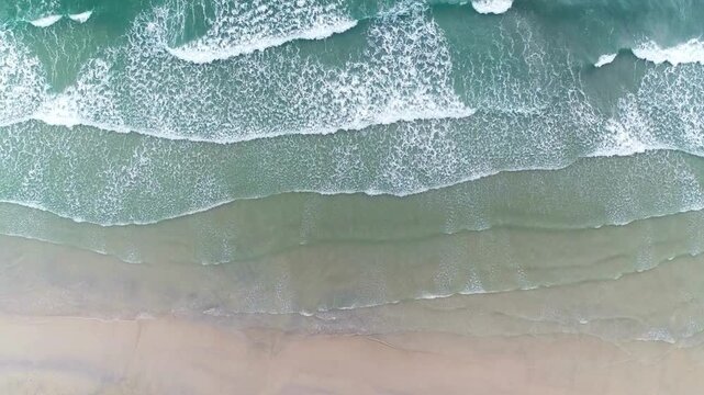 Top view of gentle ocean waves washing onto a sandy beach, calming aerial seascape with soft foam patterns, tropical shoreline mood, natural summer background, and relaxing coastal vibes