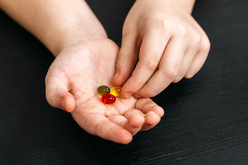 Child&rsquo;s hands holding three colorful pills in red, yellow, and green, resting on a dark wooden surface with a soft focus background. Concept medication, healthcare, pediatrics
