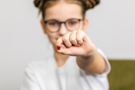 Young girl with glasses holds a small red pill in her hand, wearing a white t-shirt, with a soft-focus background of a green couch and neutral wall