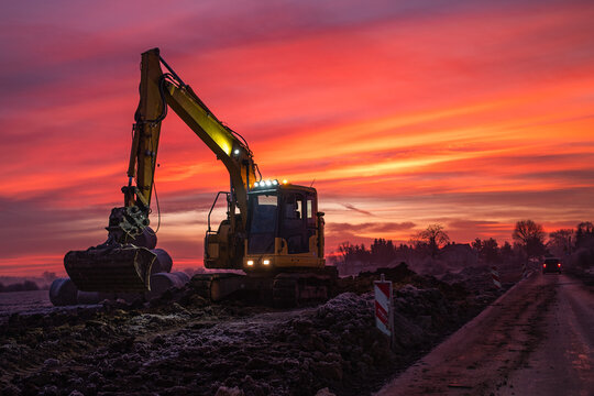 A yellow excavator stands on a construction site next to a road, silhouetted against a dramatic and vibrant orange and red sunrise.