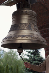 Fototapeta premium Close-up of a hand pulling a chain to ring a large, ornate bronze bell at an outdoor monument.
