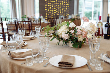 Close-up of wedding table setting with crystal glassware, plates, and beige napkins. Luxury banquet dining details with fresh floral centerpiece and bright window light